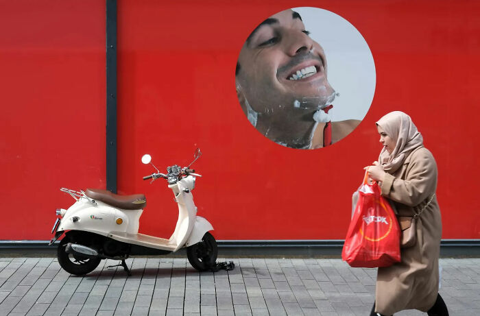 Woman walking past a scooter on a city street with a red wall featuring a large smiling face street shot.