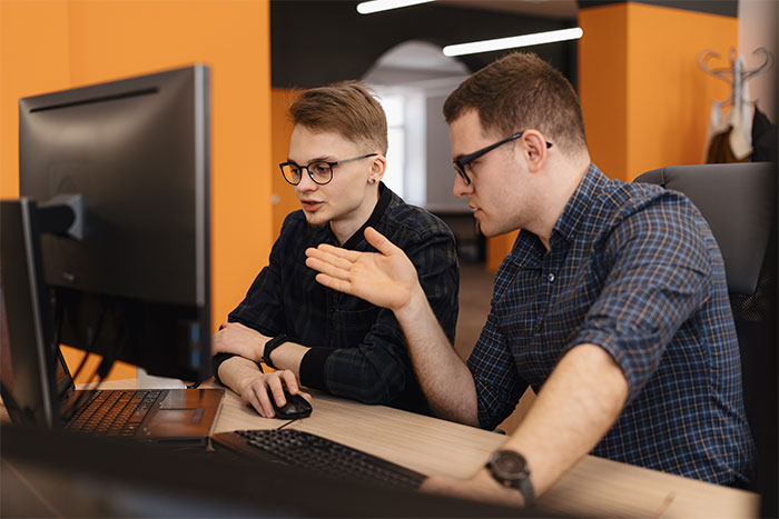 Two employees discussing deployment rule details intently while working at computers in a modern office setting. Two employees discussing deployment rule details intently while working at computers in a modern office setting.