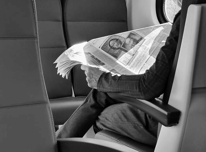 Black and white street shot of a person reading a newspaper on public transport, capturing the poetry of everyday life.