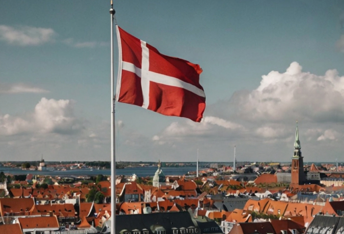 Danish flag waving over a cityscape with rooftops and a church tower, representing best countries to live in 2025. Danish flag waving over a cityscape with rooftops and a church tower, representing best countries to live in 2025.