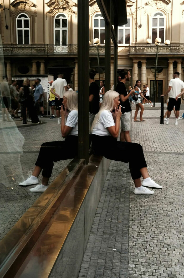 Woman sitting and smoking reflected in glass window on busy street, capturing beautiful street shots of everyday life.