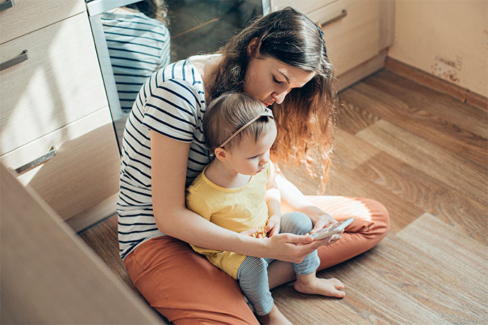 Woman babysitting toddler during family holiday, sitting on wooden floor and using a smartphone together. Woman babysitting toddler during family holiday, sitting on wooden floor and using a smartphone together.