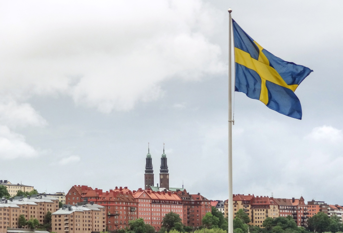 Swedish flag waving over cityscape representing best countries to live in 2025 with historic buildings and cloudy sky background Swedish flag waving over cityscape representing best countries to live in 2025 with historic buildings and cloudy sky background