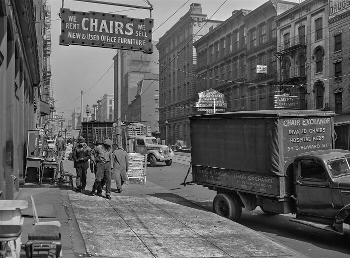 Colorized historical photo showing people and vintage trucks near a chair exchange sign on a city street.