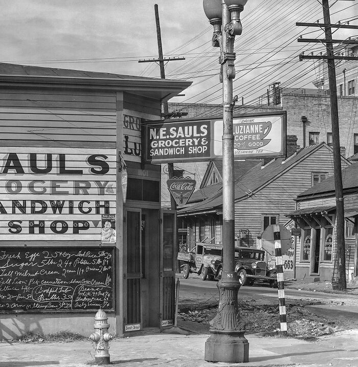 Colorized historical photo of an old grocery and sandwich shop on a street corner with vintage cars nearby.