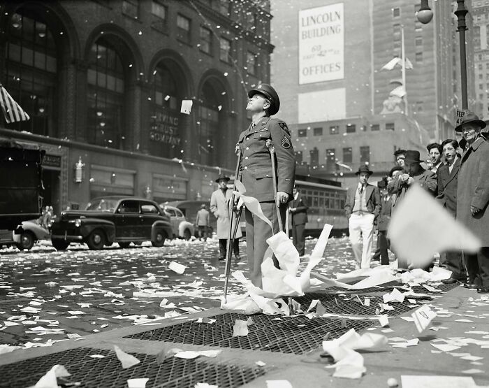 Colorized historical photo of a soldier on crutches surrounded by paper debris in a busy urban street scene.