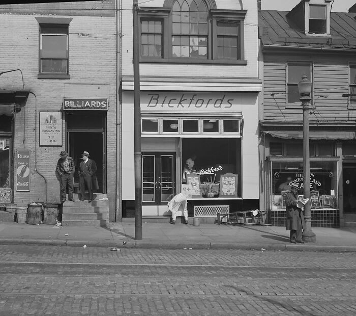 Colorized historical photo showing early 20th-century street scene with shops and people interacting outside.