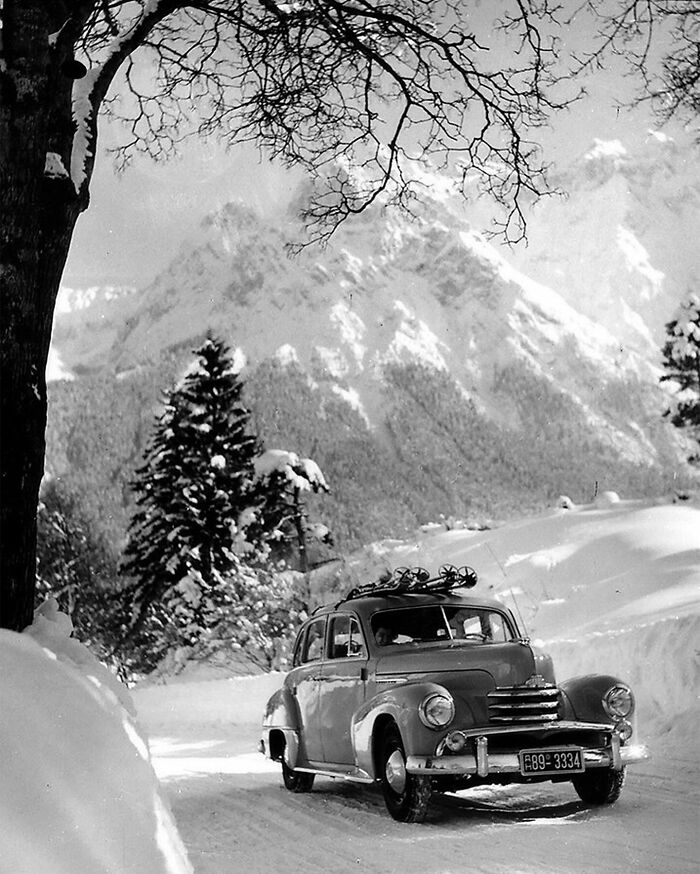 Vintage red car driving on snowy mountain road with snow-covered trees, colorized historical photo by talented viral artist