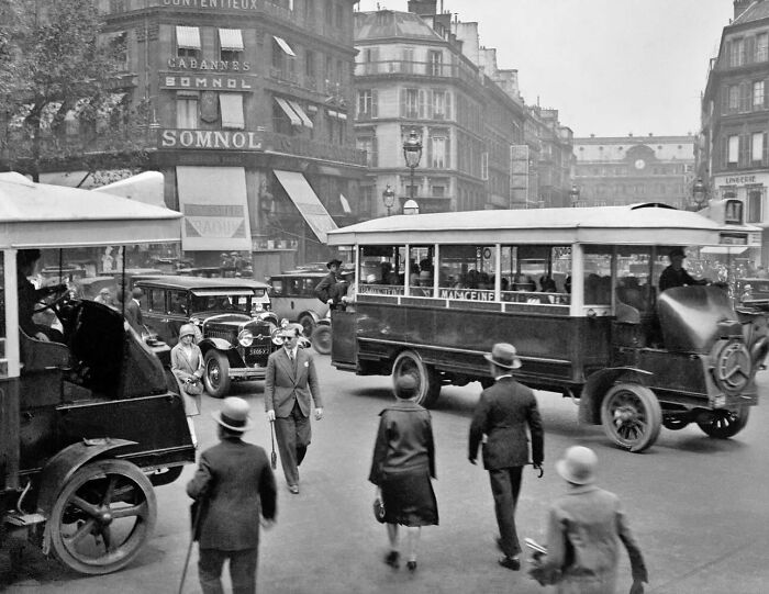 Colorized historical photo showing 1920s city street with vintage cars, tram, and people dressed in period clothing.