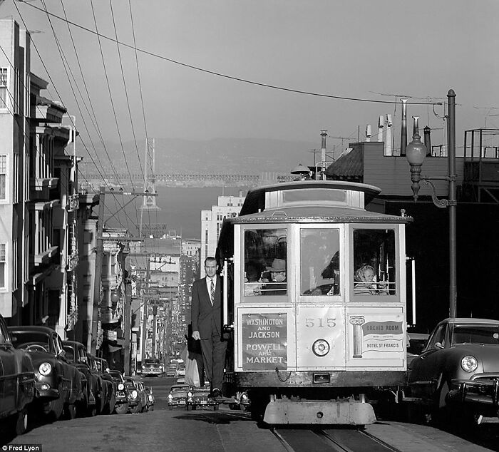 Colorized historical photo of a San Francisco cable car on a street with passengers and vintage cars in the background.