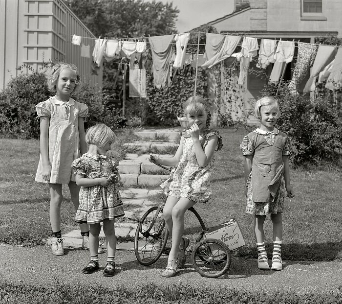 Four children in vintage dresses outside a house with colorful clothes hanging on a line, colorized historical photo.