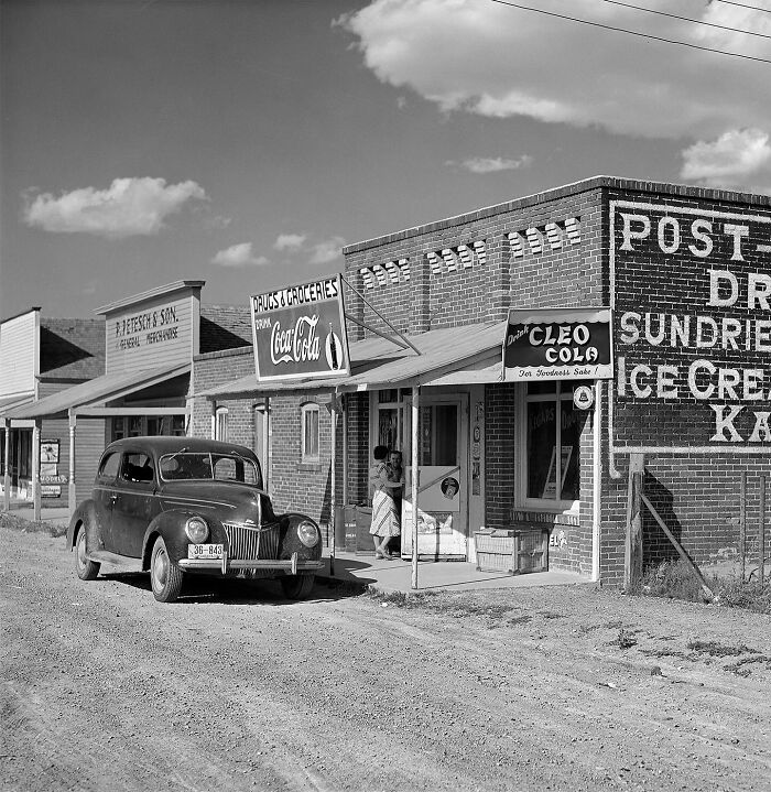 Colorized vintage photo of a rural grocery store and classic car on a dusty road from magical historical photos collection.