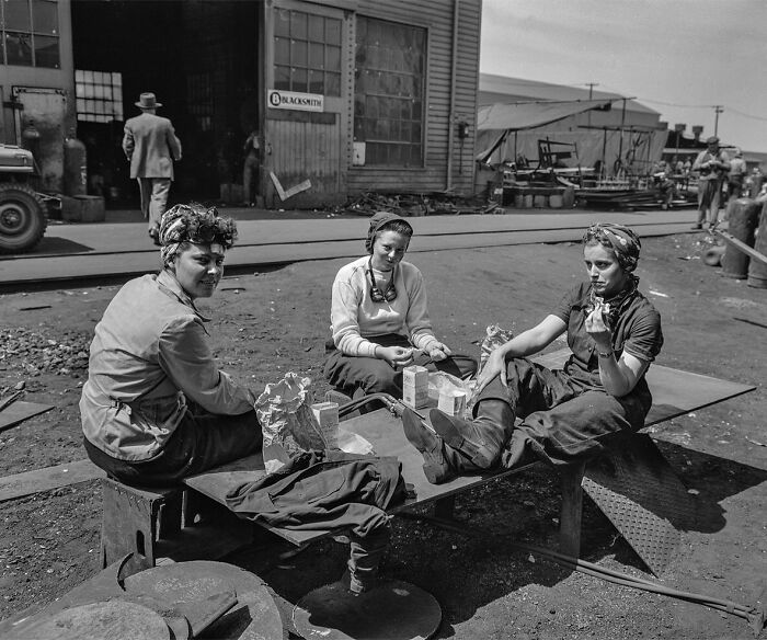 Three women on a break at a workshop, colorized historical photo showing workers in vintage clothing and industrial setting.