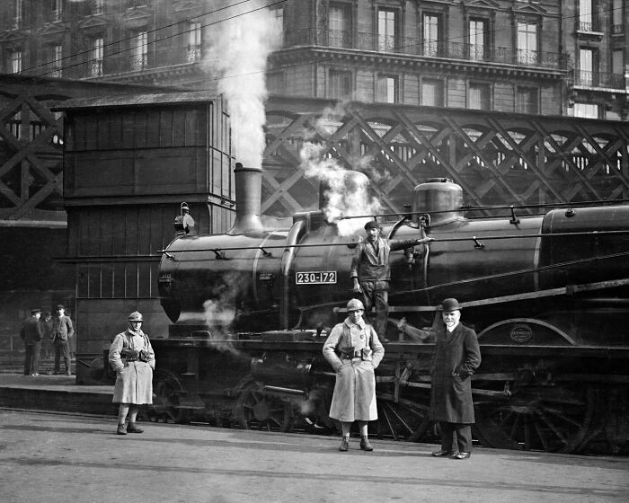 Colorized historical photo showing workers and officers near a steam locomotive at an urban train station platform.