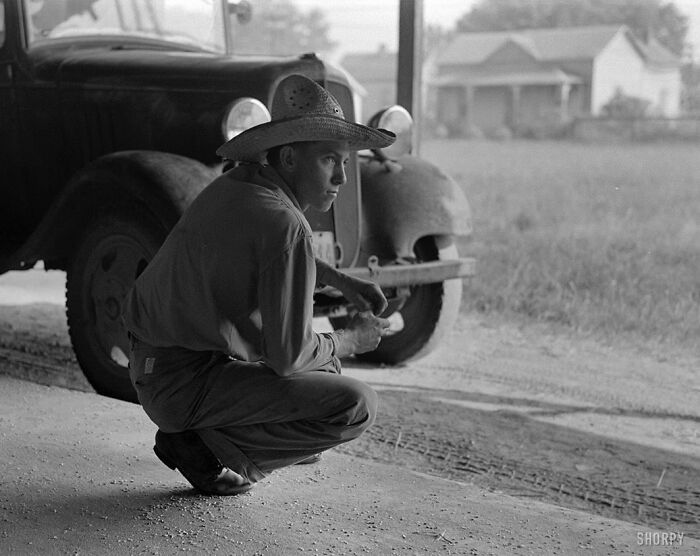 Young man in a straw hat crouching by a vintage green truck in a rural setting, colorized historical photo.