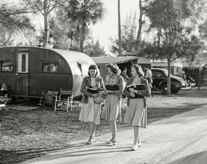 Three women in colorful vintage dresses walking with books near trailers in a magical historical photo colorized by a viral artist.