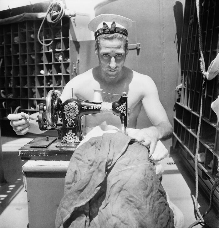 Colorized historical photo of a sailor using a vintage sewing machine to mend a large red fabric.