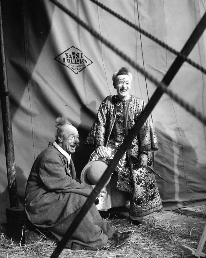 Two clowns in colorful costumes sitting and smiling against a red circus tent in colorized historical photo.