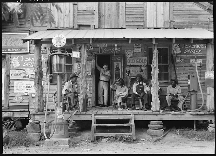 Porch scene of a vintage wooden store with men sitting and standing, colorized historical photo by a viral artist.