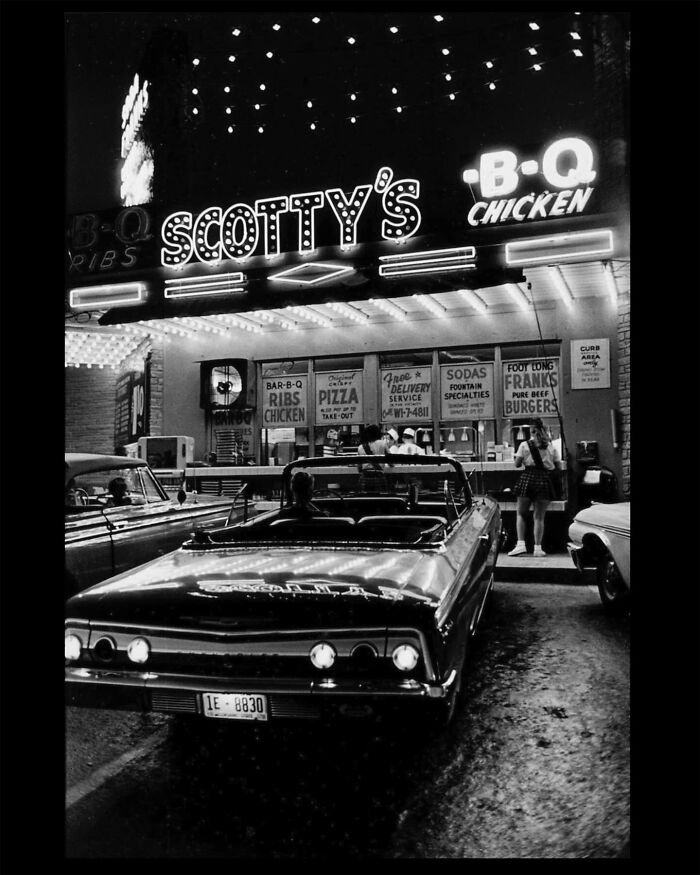 Colorized historical photo of a vintage 1960s car parked outside Scotty's BBQ chicken restaurant at night.