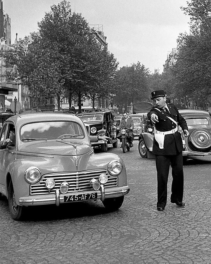 Colorized historical photo of a traffic policeman directing vintage cars on a tree-lined street.