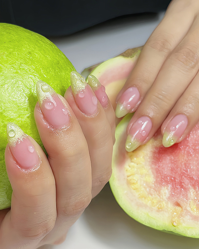 Hand with summer nails featuring glossy pink polish, yellow tips, and floral and bee 3D nail art decorations.
