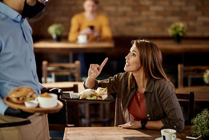 A woman speaking to a masked waiter while food is being served, showing a scene of good trend slowly disappeared.