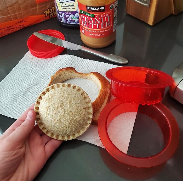 Hand holding a sealed sandwich made with a kitchen item alongside peanut butter, jelly, and a red sandwich cutter on a counter.