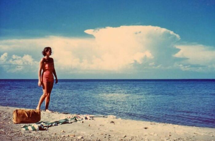 Woman in vintage swimsuit standing on a beach with a bag and towel, a mesmerizing vintage photo capturing history.