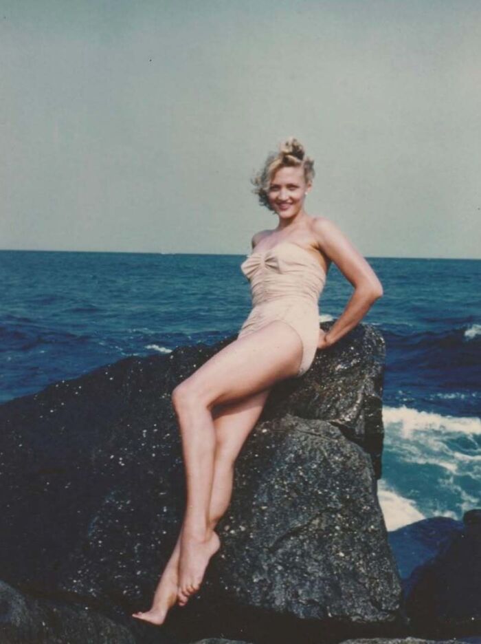 Vintage photo of a smiling woman in a swimsuit posing on rocks by the ocean, capturing a mesmerizing moment in history.