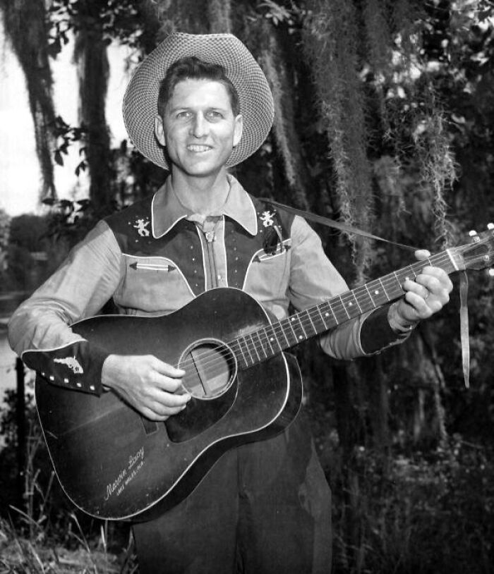 Vintage cowboy playing guitar outdoors, wearing a western shirt and cowboy hat in a classic old west setting.