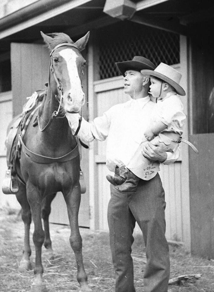 Man holding child wearing cowboy hats standing next to a saddled horse in vintage cowboy photograph.
