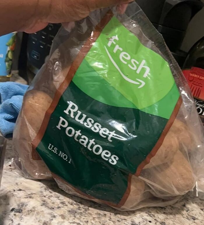 Bag of Amazon Fresh Russet Potatoes held by hand on a kitchen countertop, showcasing affordable grocery staples.
