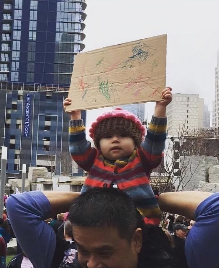 Child wearing a knitted hat holding a cardboard sign while sitting on an adult's shoulders during a crowded outdoor event.