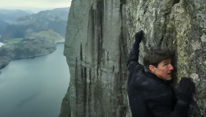 Man climbing steep cliff face overlooking river, a thrilling scene from one of the new millennium's greatest films.