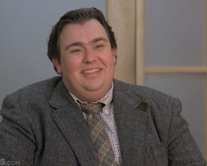 Actor smiling warmly in a gray suit and plaid tie, representing stars we lost too soon in the 20th century.