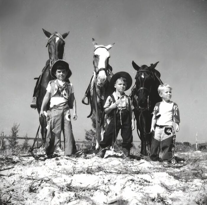 Three young boys dressed as vintage cowboys stand in front of their horses in a western outdoor setting.