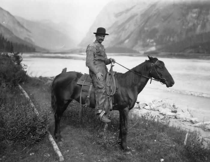Vintage cowboy wearing a hat and chaps sits on horseback near a river in a mountainous wild west landscape.