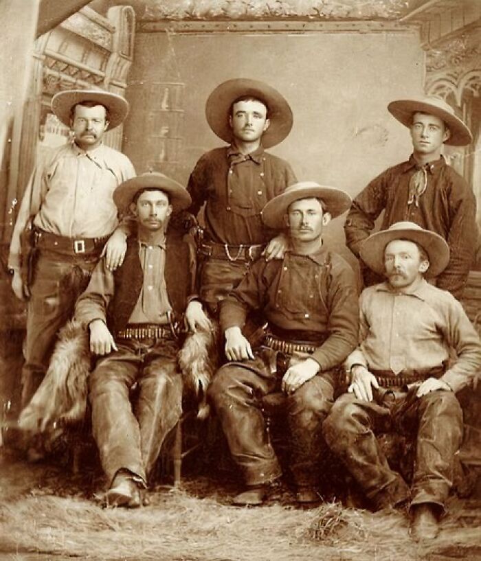 Vintage cowboy photograph showing six men in wide-brimmed hats and period attire posing in a rustic indoor setting.