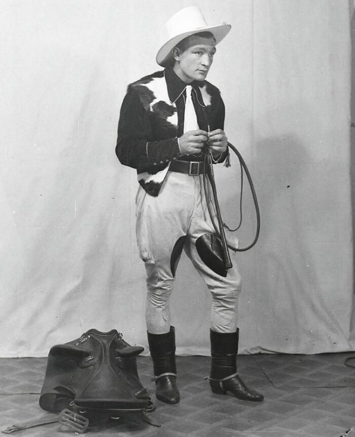Vintage cowboy dressed in Western attire holding a lasso with saddle gear on the floor in a studio setting
