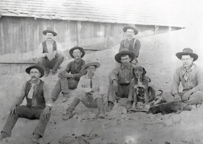 Group of vintage cowboys in hats sitting outside a wooden building with dogs, capturing the old west cowboy lifestyle.