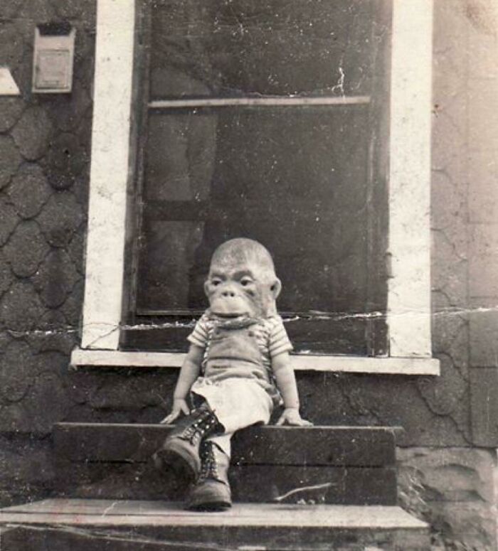 Black and white photo of a child wearing an ape mask sitting on steps, a creepy and unsettling vintage image from the past.