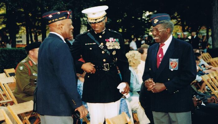 Three African American military veterans in uniform and blazers interacting outdoors at a commemorative event.