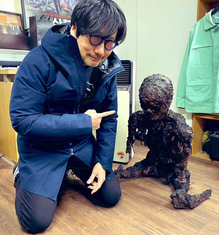 Man in glasses and blue jacket kneeling indoors next to a burnt sculpture, illustrating internet lies with absurd humor.
