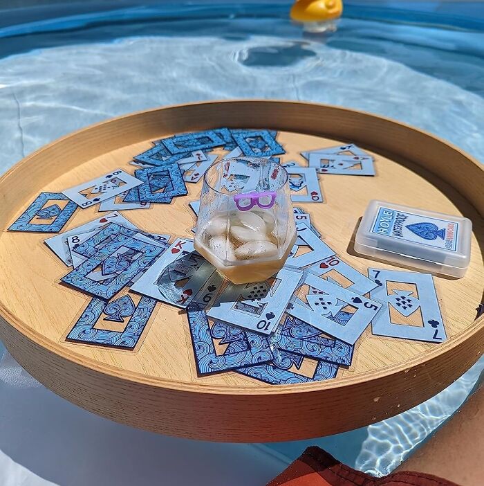 Glass of iced drink and playing cards on a wooden tray floating in a pool to fight summer annoyances.