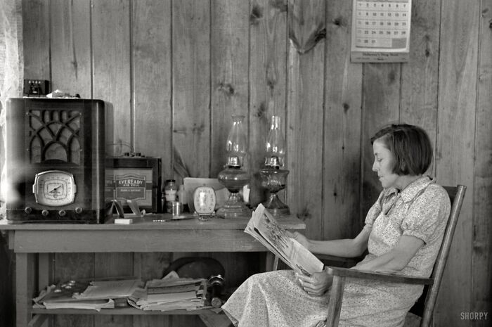 1940s housewife sitting in a wooden chair reading a newspaper in a rustic room with vintage decor and radio.