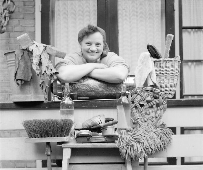 Smiling 1940s housewife leaning on kitchen counter surrounded by cleaning tools and household items in black and white.