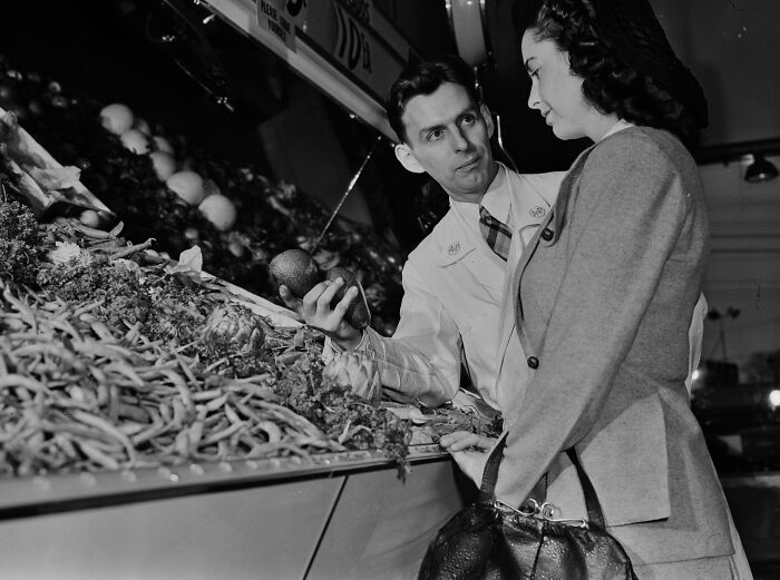 1940s housewife shopping for fresh produce assisted by a store clerk in a black and white vintage photo.