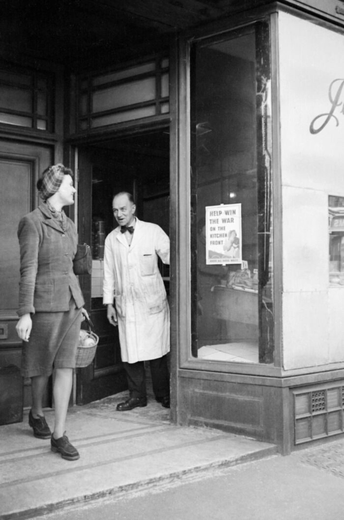 1940s housewife carrying a basket, leaving a shop with a man in a white coat standing in the doorway.