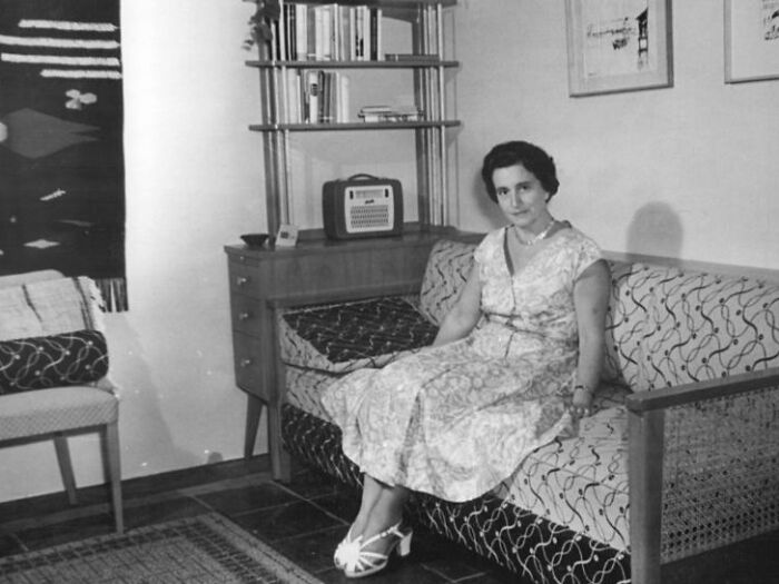 1940s housewife sitting on patterned sofa in vintage living room with bookshelf and radio in black and white photo